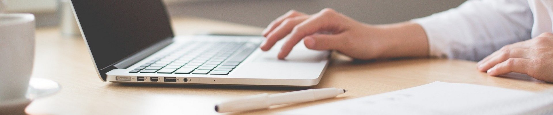 Eine Frau in einer weißen Bluse sitzt an ihrem Schreibtisch auf dem ein Block liegt und arbeitet an ihrem Laptop