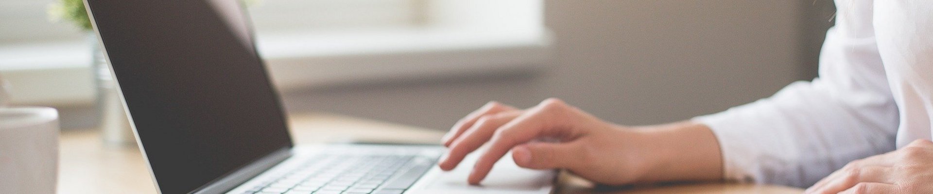 Eine Frau in einer weißen Bluse sitzt an ihrem Schreibtisch auf dem ein Block liegt und arbeitet an ihrem Laptop