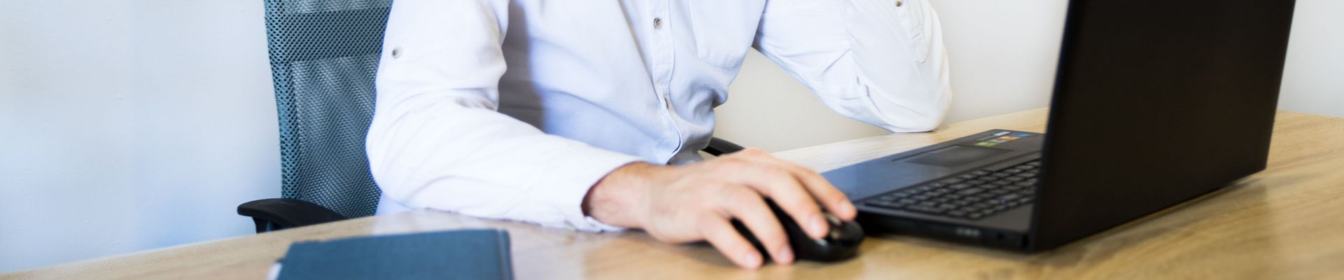 Ein Mann in einem rosa Hemd sitzt an einem Schreibtisch, lächelt und arbeitet an seinem Laptop.