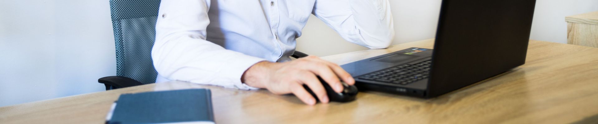 Ein Mann in einem rosa Hemd sitzt an einem Schreibtisch, lächelt und arbeitet an seinem Laptop.