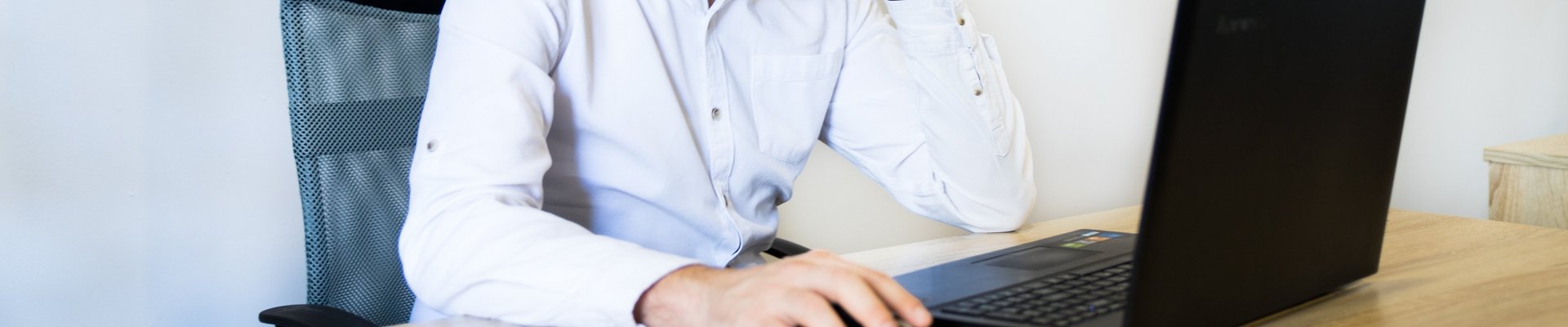Ein Mann in einem rosa Hemd sitzt an einem Schreibtisch, lächelt und arbeitet an seinem Laptop.
