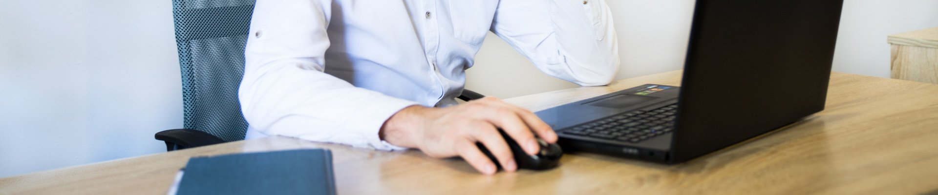 Ein Mann in einem rosa Hemd sitzt an einem Schreibtisch, lächelt und arbeitet an seinem Laptop.