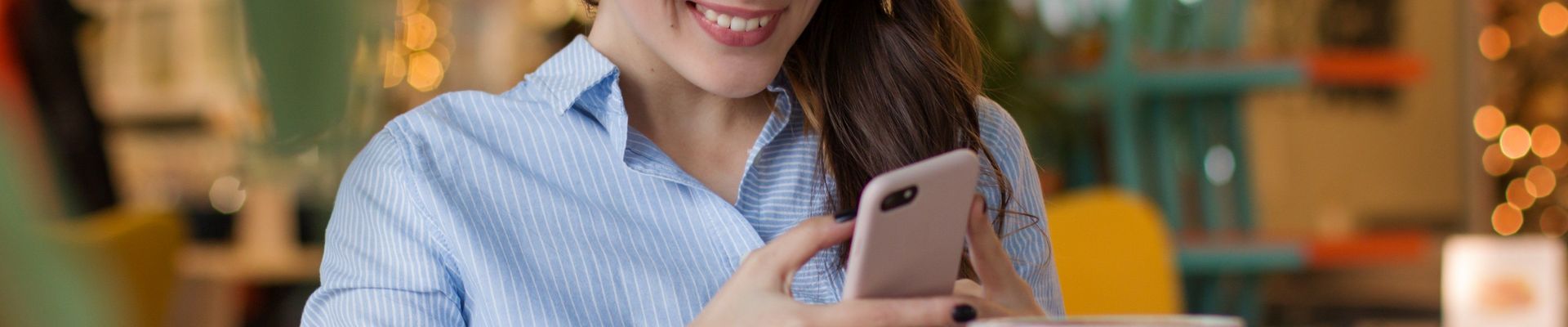 Eine Frau trinkt Kaffee, hat ihr Handy in der Hand, guckt auf den Bildschirm und lächelt.