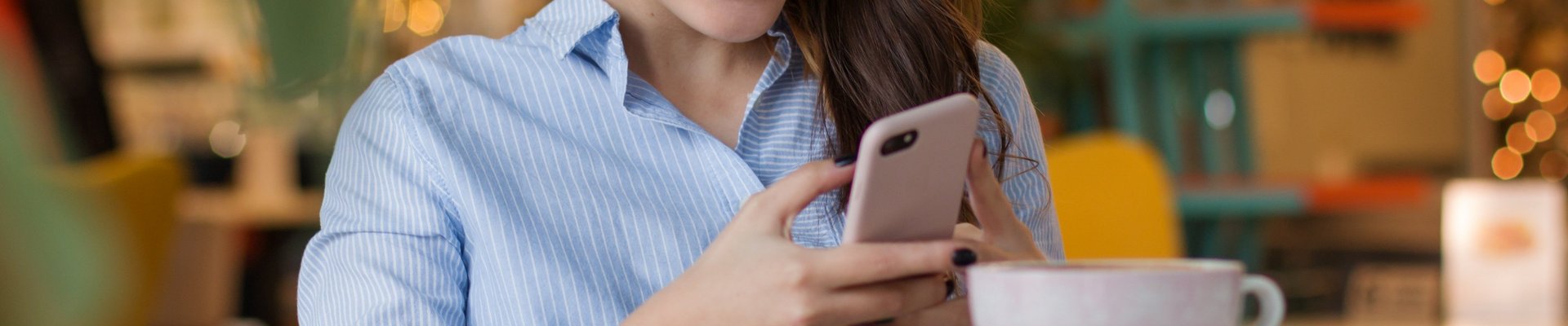Eine junge Frau sitzt in einem Kaffee an einem Tisch, ließt etwas auf ihrem Handy und lächelt