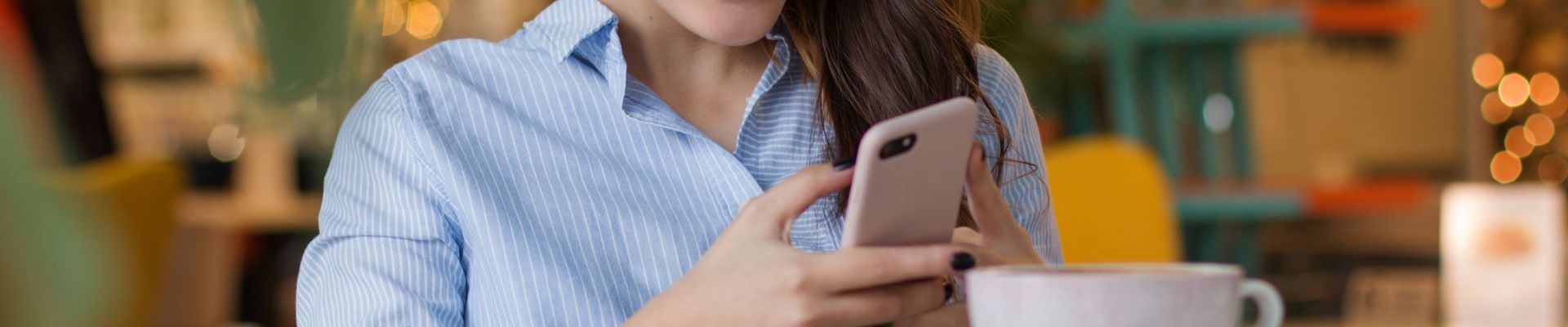 Eine junge Frau sitzt in einem Kaffee an einem Tisch, ließt etwas auf ihrem Handy und lächelt