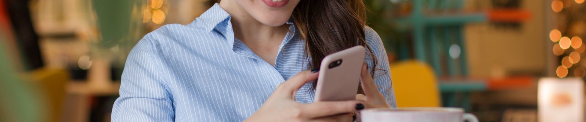 Eine junge Frau sitzt in einem Kaffee an einem Tisch, ließt etwas auf ihrem Handy und lächelt