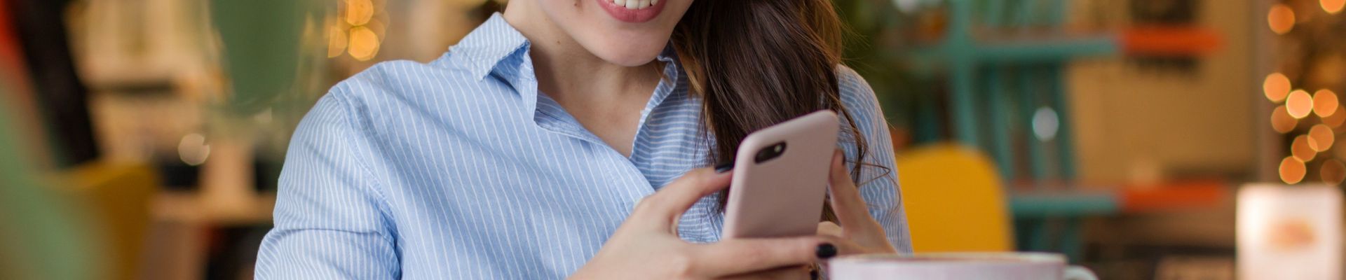 Eine junge Frau sitzt in einem Kaffee an einem Tisch, ließt etwas auf ihrem Handy und lächelt