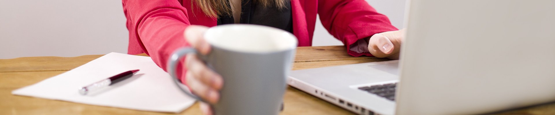 Eine Frau sitzt mit einem Kaffe am Schreibtisch und arbeitet an ihrem Laptop.
